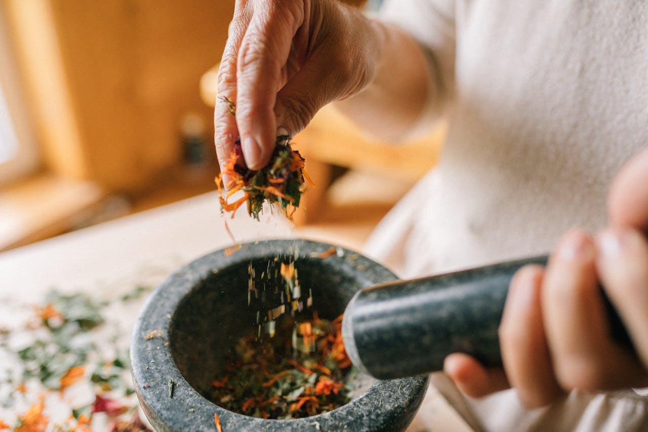 Hands using a mortar and pestle to grind dried herbs on a wooden table.